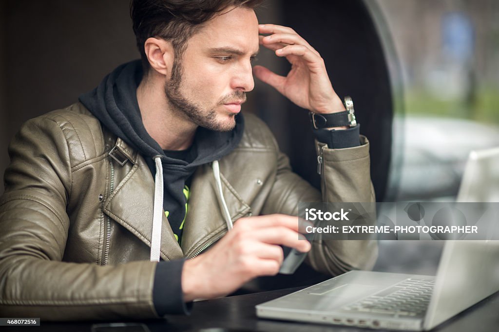 Man in coffee shop with laptop computer Young male digital nomad in the coffee shop holding cup of coffee while reading some content on his laptop computer. He is wearing leather jacket and hoodie. The photo is taken through the glass window hense the reflection on the right side. 2015 Stock Photo