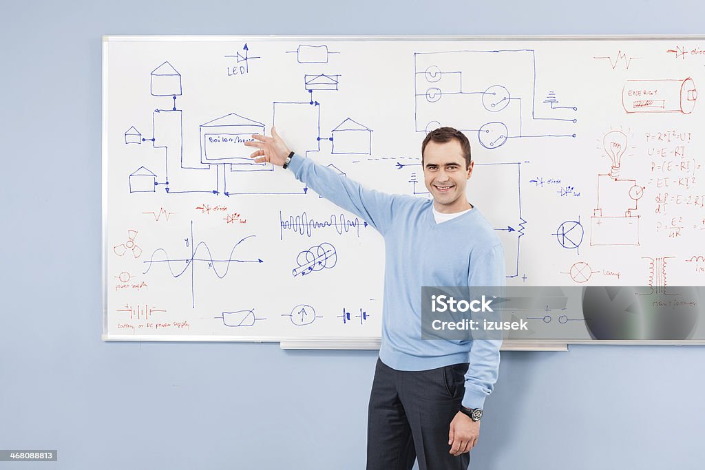 Teacher Portrait of male teacher pointing at diagrams technical systems drawn on whiteboard, smiling at the camera. Electrician Stock Photo