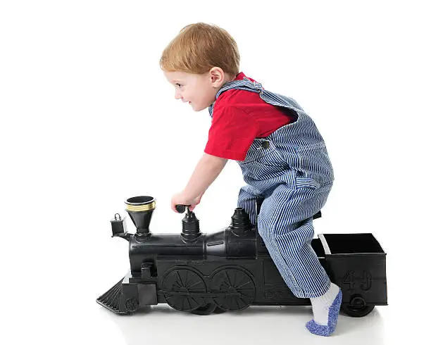 Sied view of an adorable 2-year-old train engineer scooting along on a toy train engine.  On a white background.