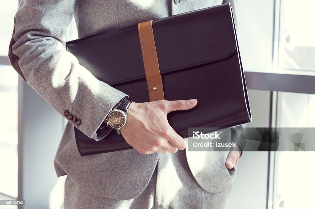 Businessman holding briefcase, close up Businessman wearing grey suit standing in an office and holding black leather briefcase. Close up of hands, unrecognizable person. Bag Stock Photo