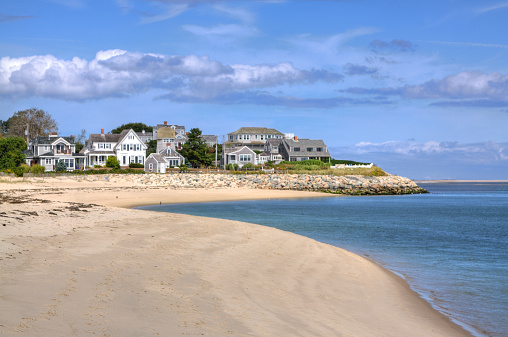 Luxury New England Waterfront Houses on a beautifu autumn day, Chatham, Cape Cod, Massachusetts. The houses are in the vicinity of Chatham Lighthouse. Sandy beach and ocean waters are in foreground. Deep blue sky with clouds is in background. HDR photorealistic image.