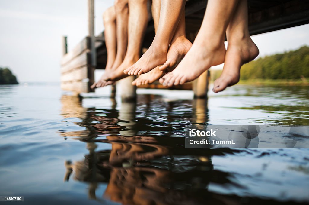 three people sitting on a jetty Legs of a bunch of young people sitting on a jetty hanging down to the water. Shot from the side during sunset. Summer Stock Photo