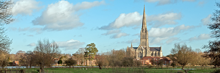 Salisbury Cathedral through trees and local buildings over the flood plains, under blue skies in Wiltshire, UK in panoramic format