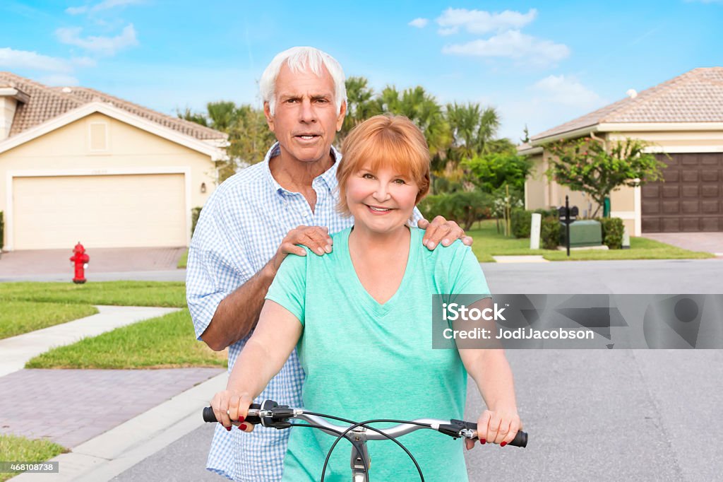 Senior couple with bicycle in residential neighborhood Caucasian senior couple posing in the street of residential neighborhood. The woman is on a bicycle and the man has his hands on her shoulders.  There are homes in back of them. It is a sunny day with a clear blue sky.  Shot with a Canon 5D Mark 3. 2015 Stock Photo