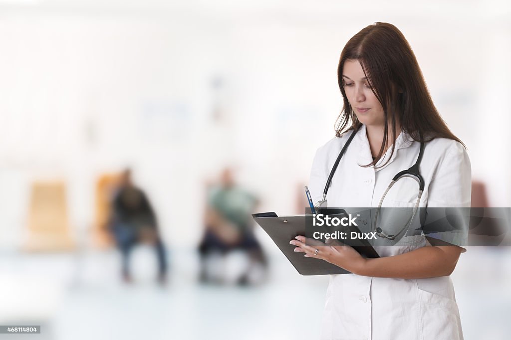 Female doctor portrait Indoor hospital portrait of beautiful female doctor with stethoscope standing and seriously writing something on clipboard. Blured background with unrecognizable patients sitting on chairs. 2015 Stock Photo