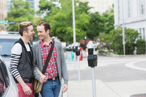 Two men, a homosexual couple, walking in the city holding hands, looking at each other, talking and smiling.  They are walking next to parked cars and a parking meter on the sidewalk.  They are wearing casual clothing.  One man is carrying a shoulder bag and wearing jeans a red plaid shirt and gray sweater.  The other is a bit more stylish, in red pants, a black vest and white shirt with black stripes on the sleeves.