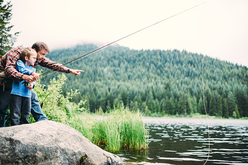 A father teaches his son how to use a fly fishing rod.  The boy focuses intently on what his dad is telling him, looking out at where the line lays in the water. Thick forest of evergreen trees visible in the background.  Shot in the beautiful Pacific Northwest at Trillium Lake, Oregon. Horizontal with copy space.