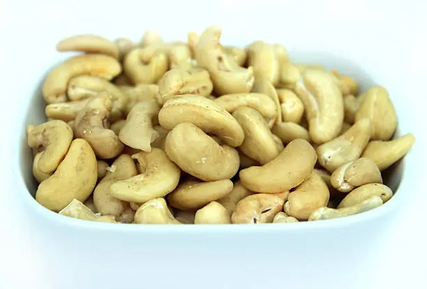 Close-up photo showing some kidney-shaped cashew nuts, which are shown shelled and piled high in a white square dish, against a white background.  Cashews are a very healthy high-protein snack food that helps to wake you up in the afternoon, while these nuts are often also used as an ingredient in salads and Chinese-style chicken stir fries.
