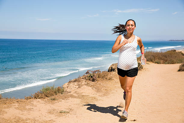 Beautiful pregnant woman jogging along the coast to stay fit A mixed race pregnant woman running on the nature trail overlooking the ocean in San Diego, California.  She is in her third trimester (7 months) and wearing a white top and maternity shorts. She's been a runner and continues to jog regularly to stay fit and strong during pregnancy. Being physically active is important for a healthy pregnancy. Copy space. 8 months pregnant stock pictures, royalty-free photos & images