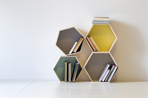 Four hexagon shaped decorative display shelves in grey, baby blue and yellow colours holding assorted books They are positioned on the floor in a disorganised fashion. the hexagons are different sizes 