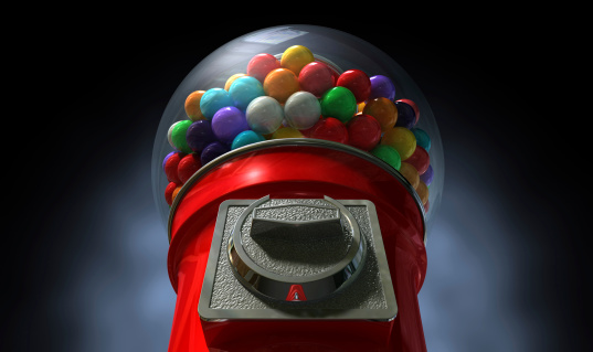 A regular red vintage gumball dispenser machine made of glass and reflective plastic with chrome trim filled with multicolored gumballs on a dark moody  background