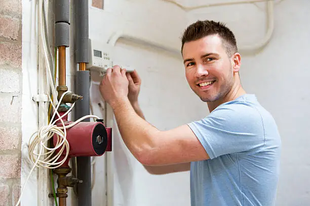 A repairman fixes a heating system while he smiles at the camera.