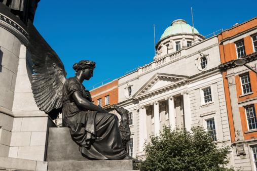 This is a horizontal, color photograph of an angel on O'Connell Street in Dublin. The statue is part of the O'Connell Monument created by sculptor John Henry Foley. Foley died prior to completion leaving his assistant, Thomas Brock to finish the monument in the late 1800s. The angel represents victory and looks towards the Neo Classical architecture that lines the thoroughfare.