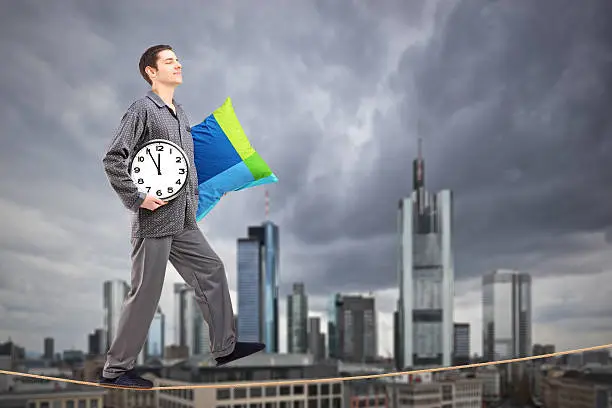 A man in pajamas holding a pillow and clock sleepwalking on a rope, with the financial centre in Frankfurt, Germany in the background