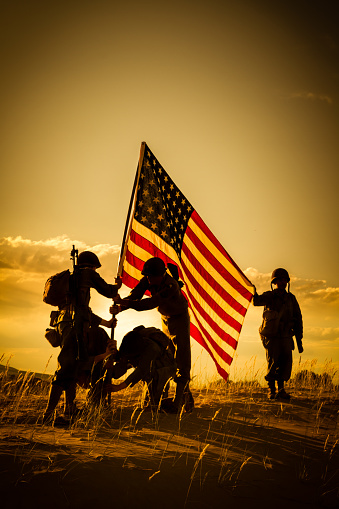 American Soldiers dressed in authentic WWII uniforms stand on a hill in the desert. They are placing the American flag into the ground. Four of the soldiers are assisting each other in standing the flag while the fifth soldier holds the flag up. There is a golden sunset behind them making the men silhouettes.
