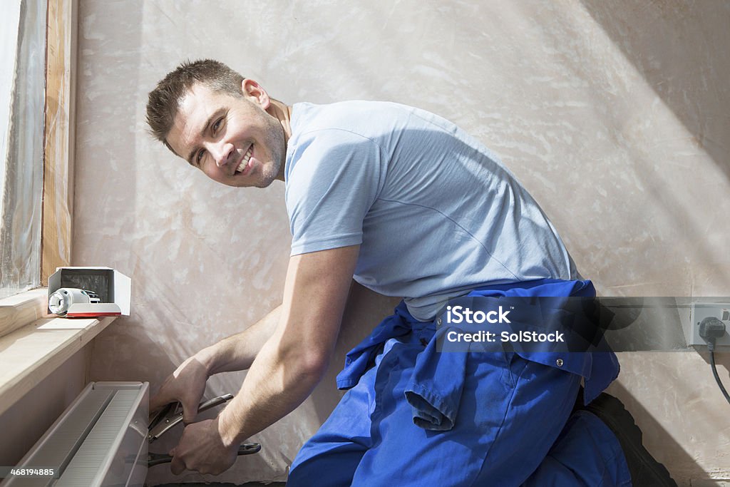 Smiley Handyman At Work A smiley handyman kneels down on the floor as he fixes a radiator in a new home. Plumber Stock Photo