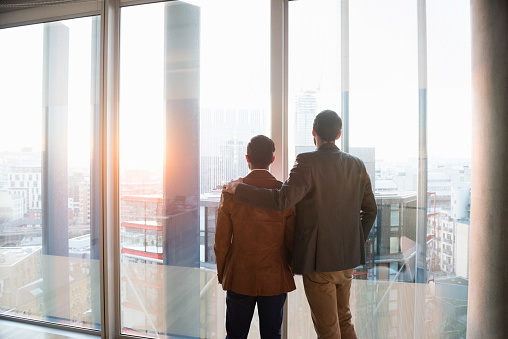 A rear view photo of businessmen standing in front of window. Professionals are in formals. Executives are admiring cityscape together, in a modern office. Two men in modern city apartment one with his arm over the others shoulder.