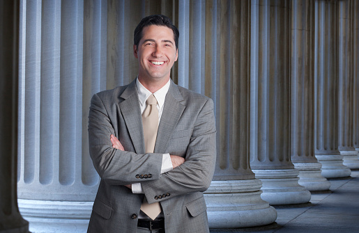 A portrait of a lawyer, banker, or businessman standing in front of a long row of columns.  He is smiling with his arms folded as he looks directly at the camera.  His jacket is unbuttoned which contributes to the casual feel of the image.  Copy space  exists on right side of image.