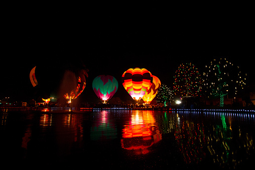 Evening hot air balloon lighting over a lake with Christmas/holiday lights decorating trees in the foreground. Night balloon firing entertains visitors during an Arizona balloon festival. Balloon reflections glow in the lake.