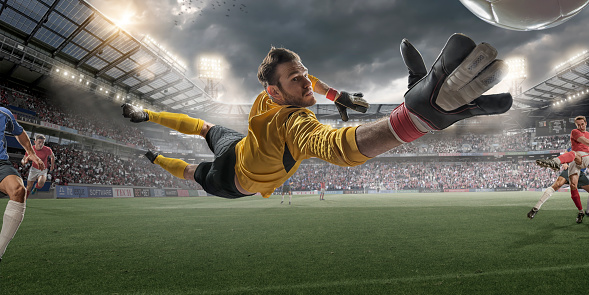 Extreme close up view from inside goal of professional soccer goalkeeper in mid air diving to save football during soccer match. The action occurs in a generic outdoor floodlit football stadium full of spectators under a stormy evening sky. All players are wearing generic unbranded kit. Stadium advertising is fake. 