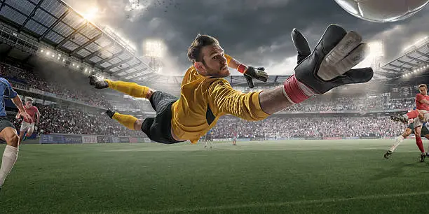 Extreme close up view from inside goal of professional soccer goalkeeper in mid air diving to save football during soccer match. The action occurs in a generic outdoor floodlit football stadium full of spectators under a stormy evening sky. All players are wearing generic unbranded kit. Stadium advertising is fake. 