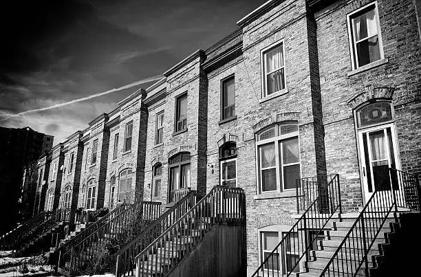 A black and white image of old brick row houses in Kitchener, Ontario. A long flight of steps leads up to the front door of the second floor apartment. Also known as townhouses in Ontario, this architectural style is fairly rare due to gentrification.