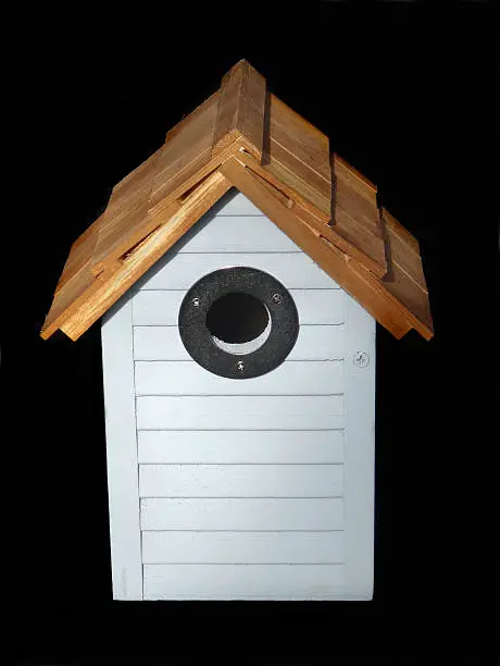 Photo showing a wooden nest box, made for small birds / finches and designed to look like a small beach hut, with a peaked roof made from wooden shingles / small cedar tiles.  A metal ring has been fixed around the entrance hole, to help prevent woodpeckers and other birds from pecking at the wood.