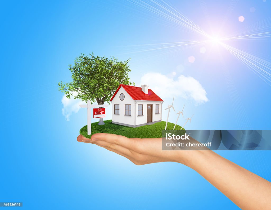 Hand holding house on green grass with red roof, chimney Hand holding house on green grass with red roof, chimney, tree, wind turbine. Near there is signboard for rent. Background clouds and blue sky 2015 Stock Photo