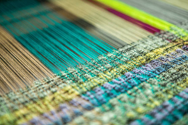 At the Loom Interwoven thread at a loom. A loom is a device used to weave cloth. The basic purpose of any loom is to hold the warp threads under tension to facilitate the interweaving of the weft threads. The precise shape of the loom and its mechanics may vary, but the basic function is the same. loom stock pictures, royalty-free photos & images