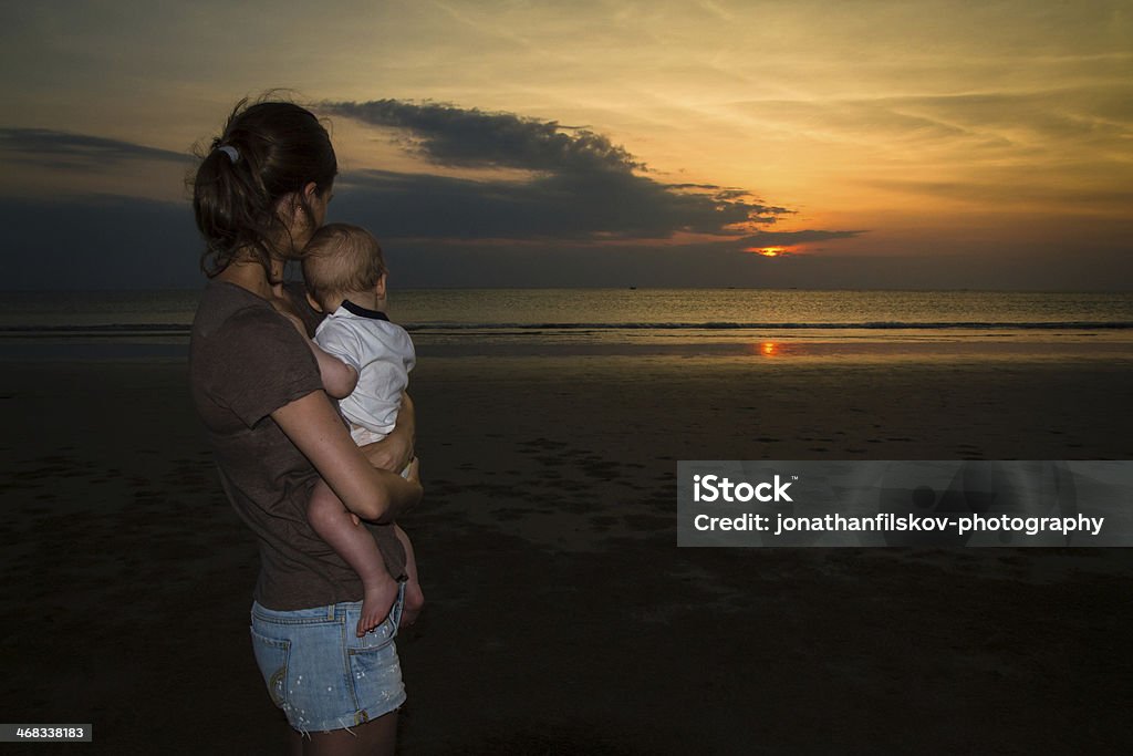 Calm waters Travelling with babies: Young mum holding her baby on ocean beach with dramatic sunset and cloudscape in background. Shot in Khao Lak, Thailand Activity Stock Photo