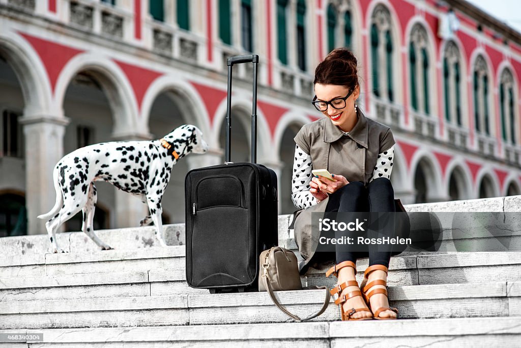 Woman sitting on outdoor steps with suitcase and Dalmatian Young woman dressed in coat and glasses with travel bag using phone on the stairs at Republic square in Split city Businesswoman Stock Photo