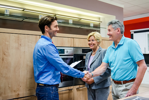 A senior couple buying a new kitchen in a modern appliance store. They finalizing their contract by a handshake with the young and smart sales clerk. Horizontal indoor shot.