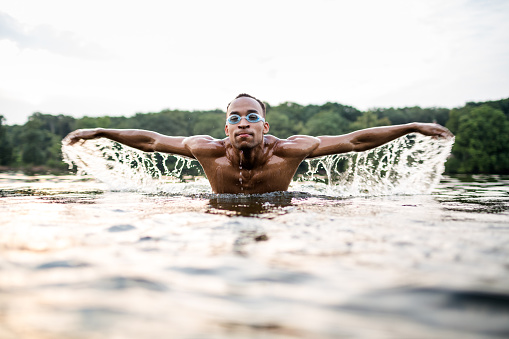 Young african man with swim goggles swimming in a lake. It's summer and he's performing butterfly swim style. Shot late evening during sunset.