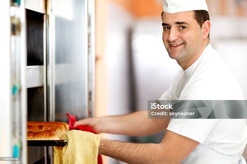 Smiling baker taking out pan of pastry from the oven. Side view of male baker taking out a pan of pastry from the oven. He is looking at the camera. Baker - Occupation Stock Photo