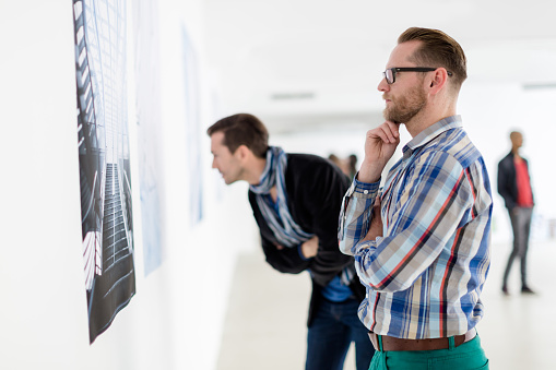 Two multi ethnic visitors of an art gallery looking at artwork hanging on the wall. Shot was taken from the artwork perspective. Focus is on young woman in the foreground looking at camera. Horizontal shot against backlit.