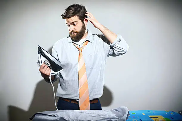 Early 30's man ironing his jacket.He's getting ready for work.He's scratching his head because he's unable to figure out how the iron is propery used.He has neat brown hair and fully grown beard.