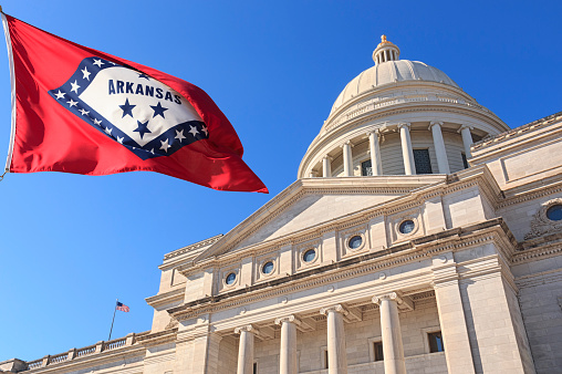 State Capitol Building front exterior in Little Rock Arkansas with US state flag outside on clear windy day with blue sky.