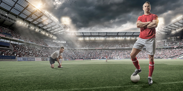 Heroic soccer player standing with foot on football and looking out into the distance with rival player bending down to tie shoelace. Players are on a soccer pitch in a generic outdoor floodlit stadium full of spectators under dramatic stormy evening sky at sunset. Players are wearing generic soccer kit.