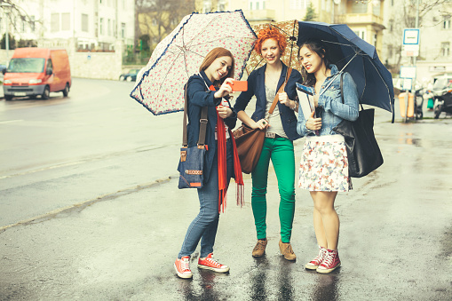 Three young women are taking selfies on a rainy day in the city. The girls are holding umbrellas. Full length shot.