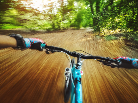 Point of view photograph of a mountain bike rider, pedaling fast in a single track immersed in the forest, with sun breaking through the right side. The background looks blurred for the high speed of the action. Picture was taken with an action cam using a chesty mount.