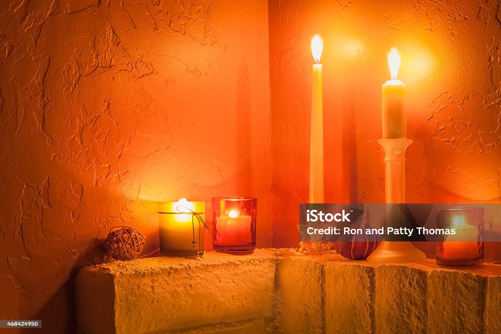 Several lit candles  on brick mantel,orange and yellow (P) Brick mantel holds two candles on candlesticks and three votive candles all lit and glowing in candlelight and glowing against rust heavily textured wall. There is copy space in the left upper quarter of image. All in rust, orange, and yellow in romantic warm colors and good year round. 2015 Stock Photo