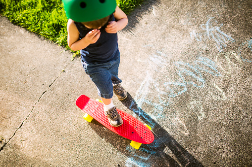 A sweet 3 year old little boy stands on a bright red skateboard with yellow wheels,  while putting on a green helmet.  