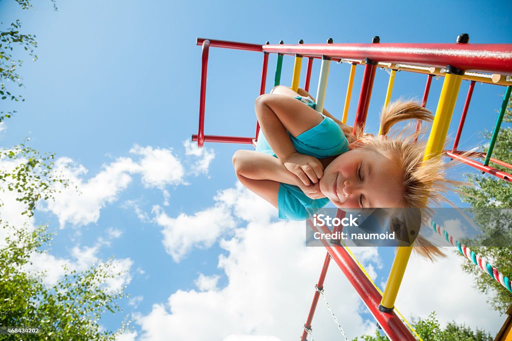 Happy child on a jungle gym Low angle view of cute blond girl wearing blue tshirt hanging from a monkey bars. Girl is smiling with her eyes closed. The climbing frame is painted in red yellow green colors and located in the courtyard of a house. Blue summer sky with clouds and tree leaves are seen in the background. Playground Stock Photo