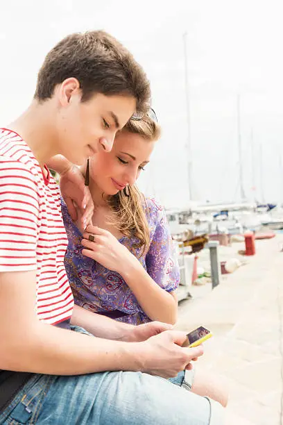 Young man of 18 in striped red and white t-shirt and shorts and woman in dress sitting tohether in front view, waist up. Woman leaning on man's shoulders and they are looking at the mobile in man's hands. In background visible some anchored boats   Summer time. 