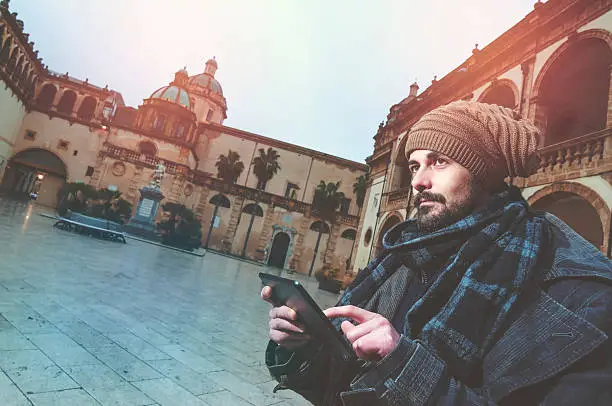 Young Man reading a tablet in a European square with a church on the background warm filter applied
