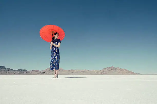 A Japanese woman wearing a Chinese style dress, stands on the Bonneville Salt Flats holding a red umbrella. Taken during the daytime with a cloudless blue sky.
