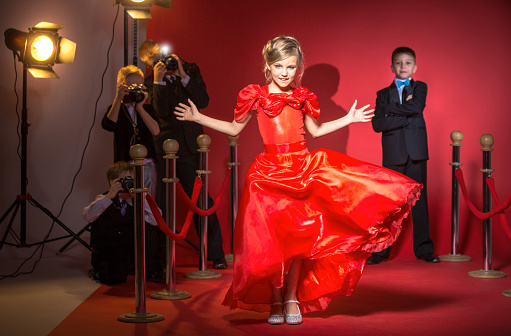 Beautiful pre-adolescent girl in red evening dress walking on a red carpet. Cute boy in a dinner suit standing behind her with his arms crossed. Group boys with photo cameras is shooting the girl celebrity