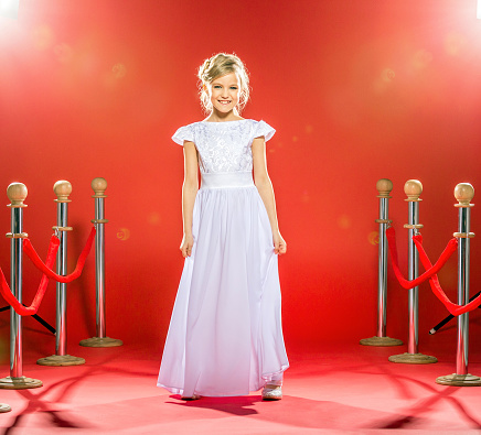Beautiful little girl in a white evening dress standing on a red carpet in the spotlight