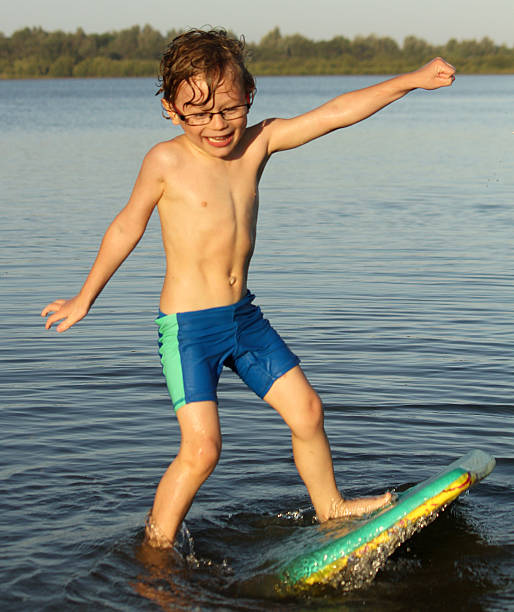 A little boy with sandy blonde hair wearing glasses, in blue and green swimmers, trying to learn how to surf by trying to stand on body board, at the river, he is happy, smiling and having fun.