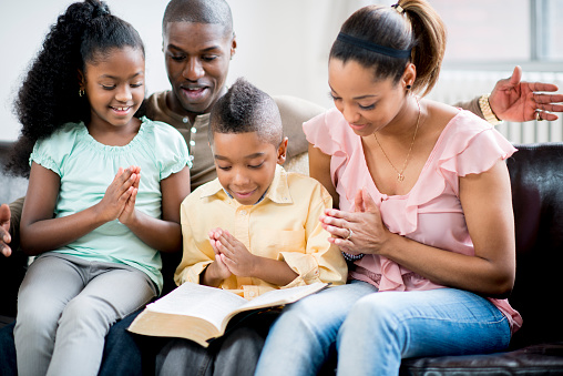 An African-American family sitting on their sofa together, folding their hands for prayer with the bible open.
